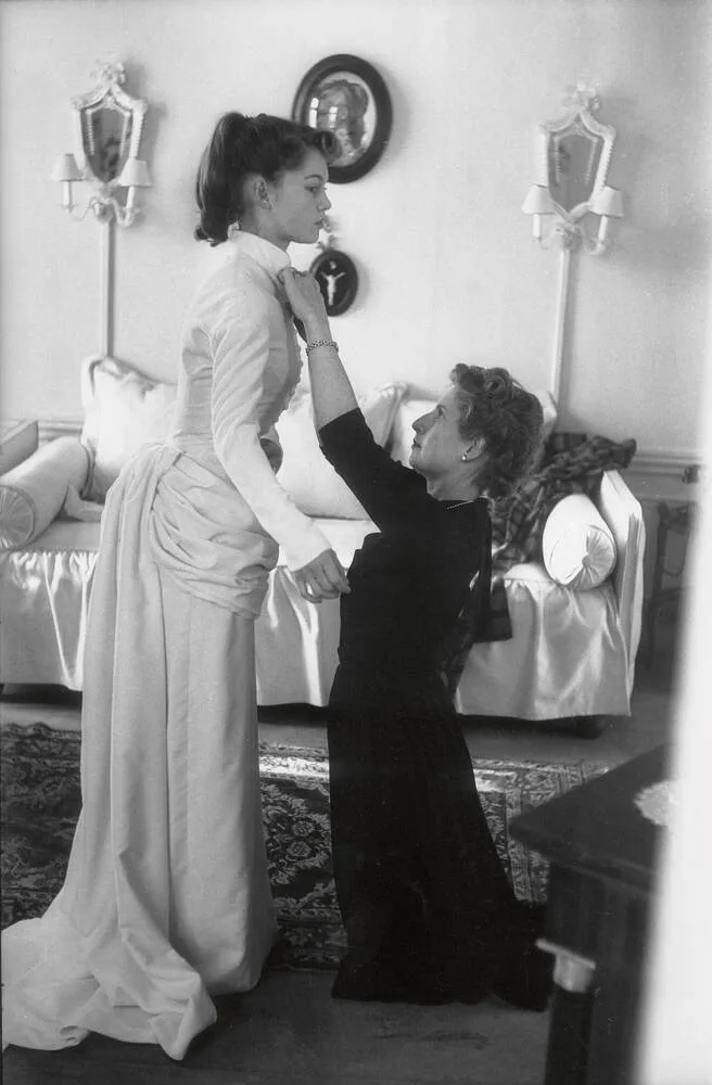 Brigitte Bardot standing in her wedding dress while a woman in a dark dress adjusts the high collar, 1952