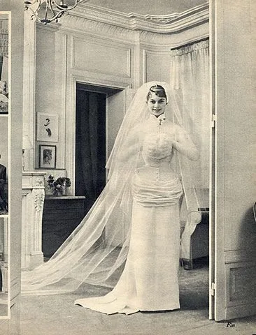 Brigitte Bardot standing in her wedding dress and long tulle veil in an apartment, 1952