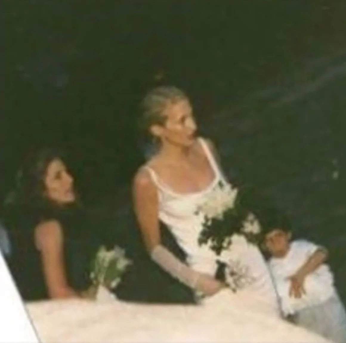 Carolyn Bessette in her wedding dress, seated with her bouquet among guests at the reception, 1996