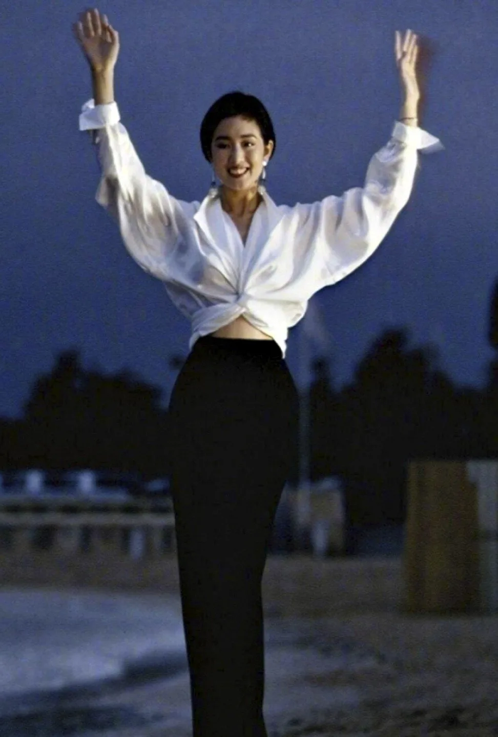 Gong Li raising both arms in a white knotted shirt on the beach at Cannes, 1993